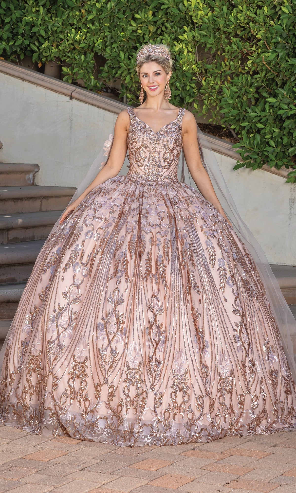 A woman smiles outdoors at the camera, wearing the Quinceanera Dress 1649 By Dancing Queen—an ornate ball gown featuring intricate embroidery, a sweetheart neckline, and a matching tiara.
